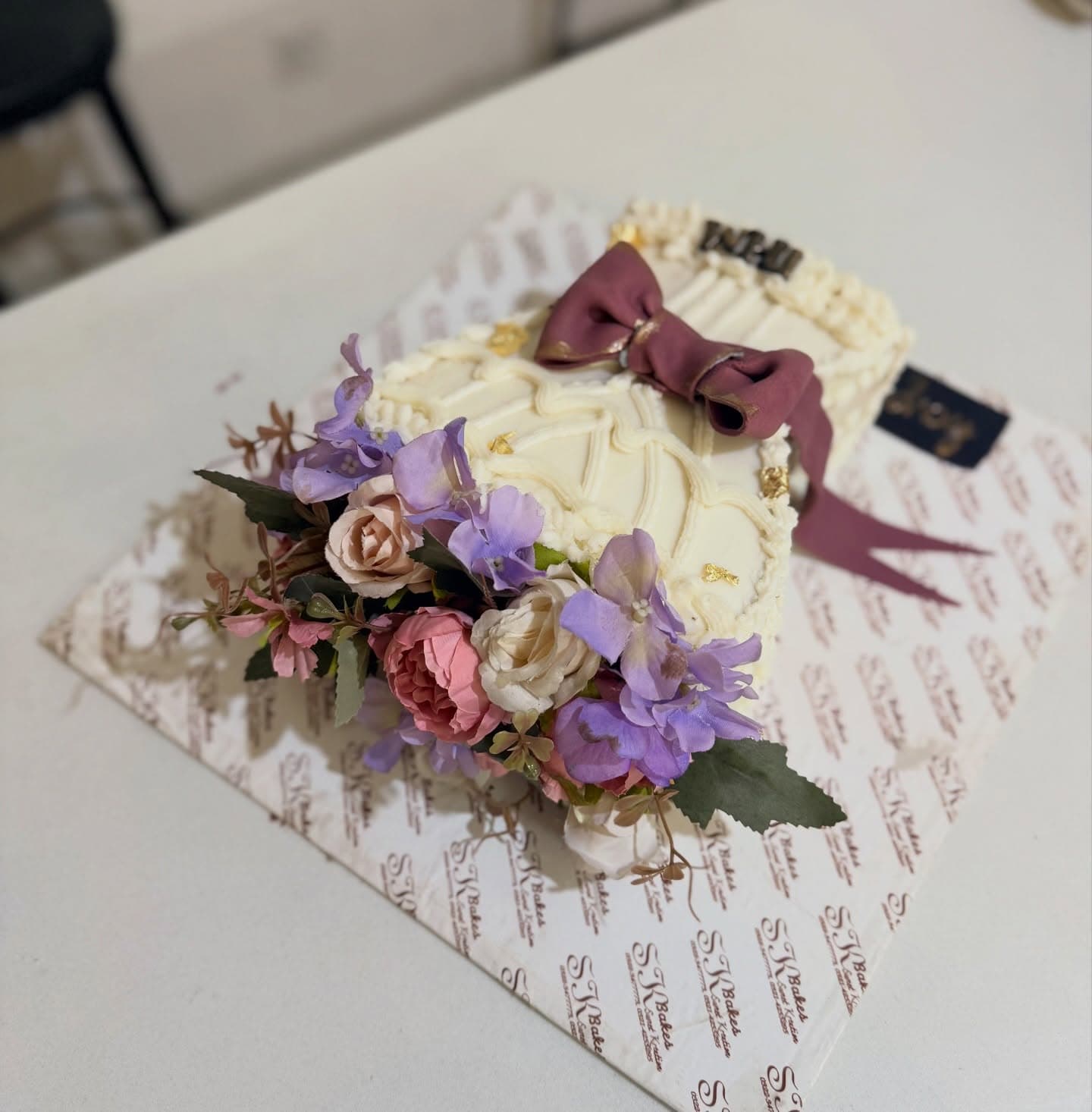 A cream-colored cake decorated with soft purple and pink flowers and a mauve bow.