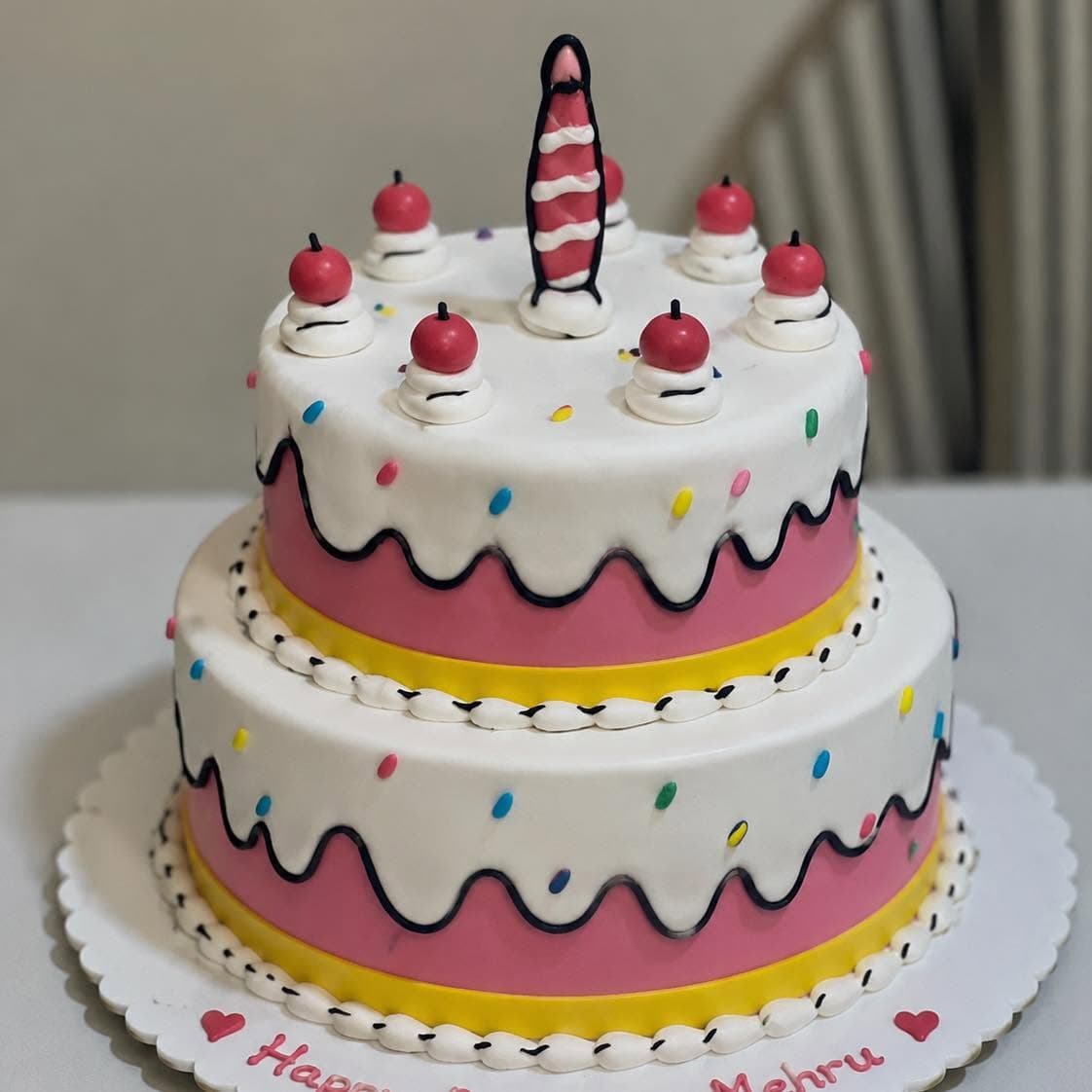 A two-tier cake with pink and white panels, dark outlines, red decorations, and a striped candle.